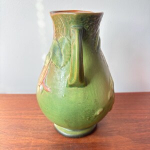 May include: Green ceramic jug with a handle and embossed floral design. The jug has a rounded body and a flared rim. The colours include shades of green, brown, and orange. The jug is sitting on a wooden surface.