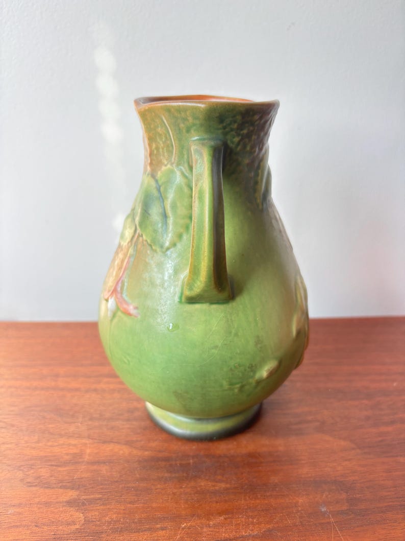 May include: Green ceramic jug with a handle and embossed floral design. The jug has a rounded body and a flared rim. The colours include shades of green, brown, and orange. The jug is sitting on a wooden surface.