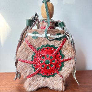 May include: A decorative flask with a wooden stopper and a fur-like exterior. The flask is adorned with red and green leather accents, including a floral design and braided details. Green and white leather straps are attached.