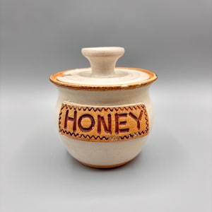 Pode incluir: Um pequeno pote de mel de cerâmica esbranquiçada com tampa e pega. A palavra "HONEY" é exibida em um rótulo decorativo marrom na frente. A borda do pote e da tampa são aparadas em marrom.