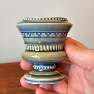 May include: A ceramic, pedestal-style vase in shades of blue and green. The vase features a decorative band of shamrocks and a beaded rim. The base is a darker green, and the top is a lighter blue. The vase is held in a hand.