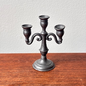 May include: A dark gray metal candelabra with three candle holders. The candelabra has a round base and ornate, curved arms. The candle holders are small and cup-shaped. The overall design is vintage and decorative.