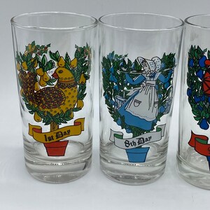 May include: Set of three clear drinking glasses with colorful illustrations. One glass features a yellow bird, another a woman in blue, and the third a red and blue design. Each glass has text on a banner.