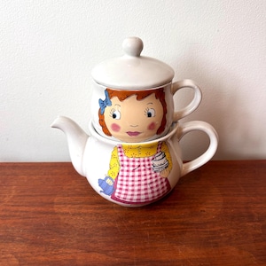 May include: A ceramic tea-for-one set featuring a white teapot base and a matching cup with a lid. The teapot displays a figure in a yellow shirt and pink checkered apron holding a blue kettle. The cup shows a face with brown hair and a blue bow.