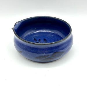May include: A small, blue ceramic bowl with a black rim and leaf design. The bowl has a slight pour spout on one side.