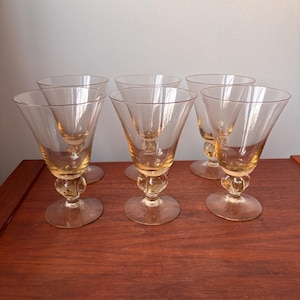 May include: Set of six vintage amber-tinted stemmed glasses. Each glass features a conical bowl, a decorative ball-shaped stem, and a circular base. The glasses are arranged on a dark wooden surface, showcasing their elegant design.