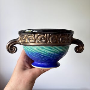 May include: A ceramic bowl with a black rim, bronze-coloured handles, and a decorative band. The bowl's body transitions from turquoise to deep blue. The handles have a curled design.