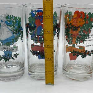 May include: Three clear glass tumblers with colorful illustrations and text from the Twelve Days of Christmas carol. The illustrations include figures and text such as "Nine Drummers Drumming" and "Ten Pipers Piping". The glasses are approximately 5 inches tall.