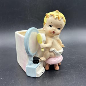 May include: A ceramic planter shaped like a baby girl sitting on a stool, holding a hand mirror and looking into it. The planter is painted in shades of pink, blue, and white.