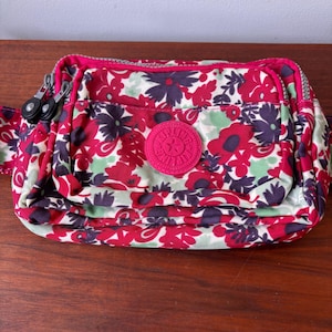 May include: A floral print crossbody bag with a pink zipper and trim. The bag features a vibrant pattern of red, purple, and green flowers on a white background. A round pink logo is centered on the front.