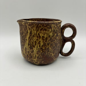 Puede incluir: Una jarra de cerámica marrón y amarilla con un asa única de doble bucle.