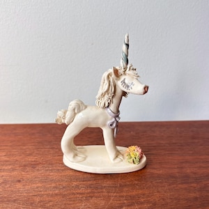 May include: A cream-coloured unicorn figurine with a spiralled horn in shades of blue and green. The unicorn has a lavender bow around its neck and a small cluster of flowers at its feet. The figurine is on a cream-coloured base.