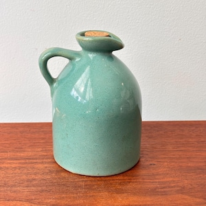 May include: A small, turquoise ceramic jug with a handle and a spout. The jug has a cork stopper and sits on a wooden surface. The jug is a vintage piece, likely used for holding liquids.