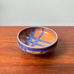 May include: A small, round ceramic bowl with an orange interior and a blue and orange geometric pattern. The rim is a deep blue colour. The bowl is on a wooden surface.