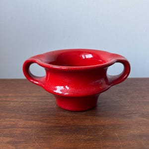 Puede incluir: Un cuenco de cerámica de color rojo brillante con un acabado liso y lustroso y dos asas laterales integradas. El cuenco tiene una abertura ancha que se estrecha hacia una base circular más pequeña, creando una silueta de color sólido.