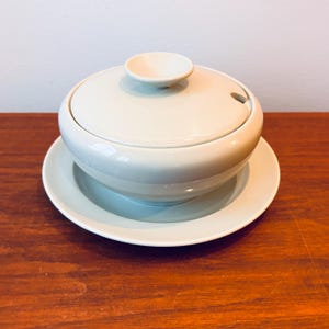 Puede incluir: Un cuenco de sopa de cerámica verde claro con tapa y un pequeño asa. El cuenco se apoya en un platillo a juego. La tapa tiene una pequeña abertura para una cuchara. El conjunto es de color verde suave y apagado.