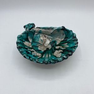 Op de afbeelding: Een teal en witte keramische schaal met een gegolfde rand. De schaal is gevormd als een schelp en heeft een getextureerd oppervlak.