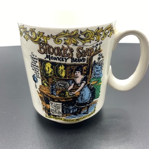 May include: White ceramic mug with a colorful illustration of a woman in a shop with the text "Brooke's Soap Monkey Brand" and a description of the soap's benefits.