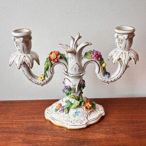 May include: A white porcelain candelabra with two arms, decorated with colourful floral accents. The base and arms have gold trim. The candelabra features a central floral design and two candle holders.