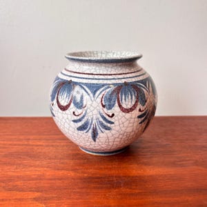 Puede incluir: Un jarrón redondo de cerámica con acabado craquelado. El jarrón presenta un diseño de patrones florales azules y granates sobre un fondo blanco. Tiene una abertura ancha y se apoya sobre una superficie de madera.