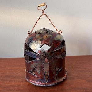 May include: A ceramic lantern with a copper wire handle and a wooden grip. The lantern has a bell shape with cut-out designs and a mottled red, blue, and gold glaze. The lantern is sitting on a wooden surface.