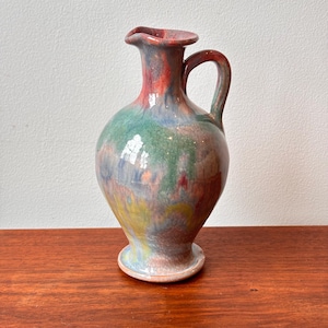 May include: A ceramic pitcher with a handle and a flared rim, featuring a blend of red, green, blue, and yellow hues. The base has a small, circular foot. The pitcher is displayed on a wooden surface. The pitcher is approximately 20 cm tall.