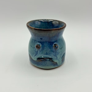 May include: A small, blue ceramic mug with a face. The mug has a glazed finish and a slightly rounded shape.