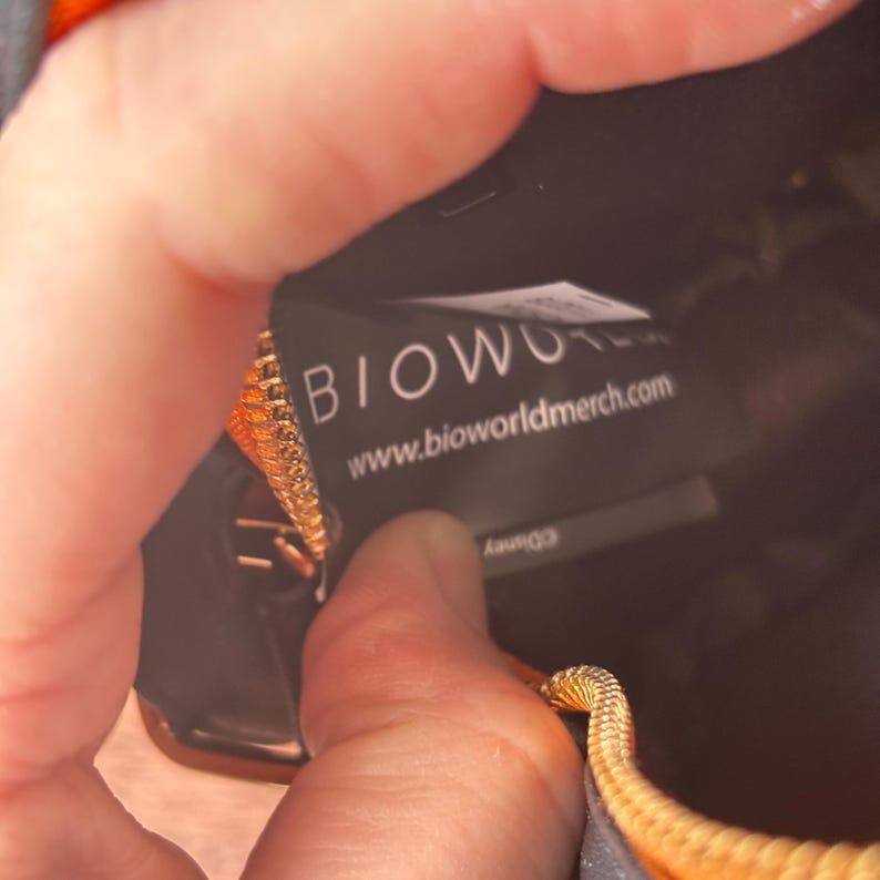 May include: Close-up of a black bag with a gold zipper. A tag inside the bag reads "BIOWORLD" and "www.bioworldmerch.com". The bag is partially open, revealing the interior.