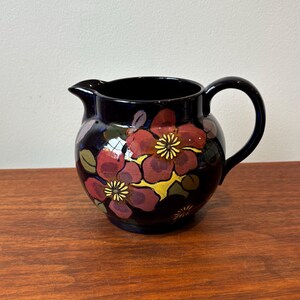 Op de afbeelding: Een donkerblauwe keramische kan met een afgeronde body en een handvat. De kan is versierd met grote, gestileerde rode en paarse bloemen met gele centra en groene bladeren. De kan staat op een houten oppervlak.