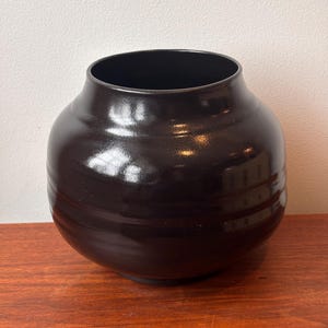 May include: A dark brown ceramic vase with a wide, rounded body and a narrow neck. The vase has a glossy finish and horizontal ridges around the body. It sits on a wooden surface.