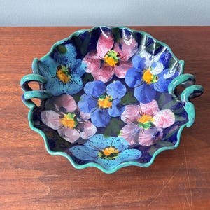 May include: A hand-painted ceramic serving dish with a floral design. The dish has a scalloped edge and two handles. The flowers are in shades of blue, pink, and yellow, set against a dark blue background. The rim and handles are teal green.