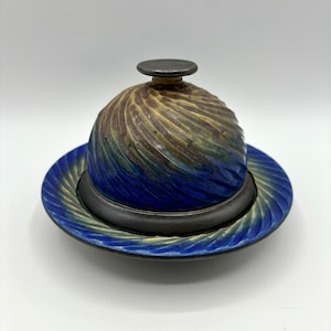 Puede incluir: Un recipiente para mantequilla de cerámica azul y marrón con un diseño de espiral. El recipiente tiene una tapa con un pequeño pomo en la parte superior.