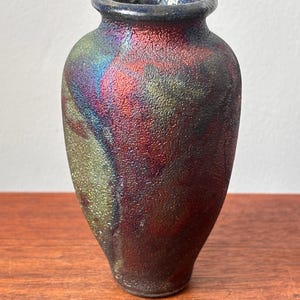 Puede incluir: Un pequeño jarrón de cerámica iridiscente con una superficie texturizada. El jarrón presenta un borde azul oscuro y un cuerpo con colores arremolinados de rojo, verde y morado. El jarrón mide aproximadamente 15 cm de alto.