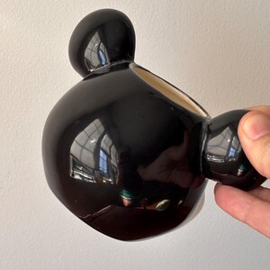 May include: A glossy black ceramic vase with a unique, rounded shape. The vase has a large, bulbous body with a smaller, spherical handle and a rounded top. The interior is a light beige color. The vase is held by a hand against a white background.