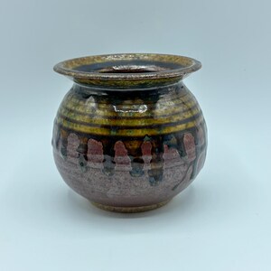 Op de afbeelding: Een kleine, ronde, bruine keramische vaas met een brede opening en een decoratief ontwerp van rode, gele en bruine strepen en druppels.