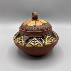 May include: A brown ceramic jar with a lid, featuring a floral design in orange, yellow, and brown. The jar's body is decorated with geometric patterns in yellow and black. The lid has a small, curled handle. A decorative storage pot.