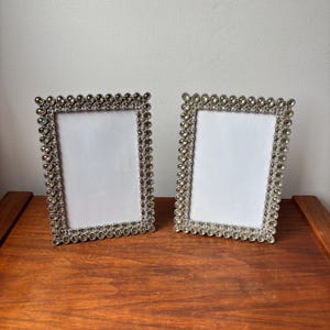 May include: Two rectangular picture frames featuring a metallic silver border encrusted with multiple rows of small, clear, oval-shaped faceted crystals. Each frame has a plain white rectangular insert centred within the decorative crystal border.