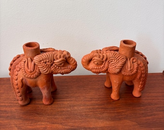 May include: Two terracotta elephant figurines with a cylindrical opening on their backs. The elephants are intricately detailed with carved patterns and stand on four legs. The figurines are a warm reddish-brown color.