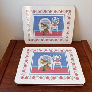 Puede incluir: Dos manteles individuales rectangulares con un diseño decorativo. Cada uno presenta una imagen central de un gato en una silla de mimbre, con un borde floral. El fondo incluye una pared azul con puntos blancos, un suelo rojo y una pequeña mesa blanca.