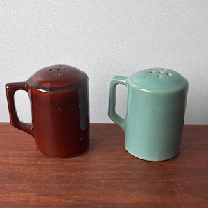 May include: A pair of ceramic salt and pepper shakers. One is a deep brown color, and the other is a light teal. Both have a handle and a lid with several holes. The shakers are on a wooden surface.