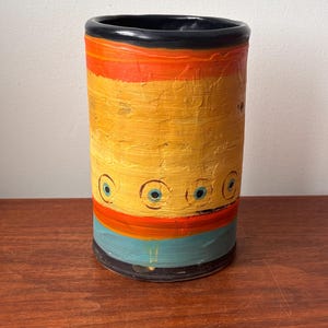 May include: A cylindrical ceramic vase with a textured surface. The vase has a black rim, with bands of orange, yellow, and teal. The yellow band has blue eye-like designs. The vase is on a wooden surface.
