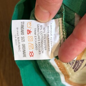 May include: A white tag with black text and care instructions. The tag says "Standard Size Ordinaire" and "Fabrique au Pakistan Made in Pakistan 50% Polyester 50% Cotton/Coton Garnitures Excluses Trim Excluded" with care instructions for washing and ironing. The tag is attached to a green and white striped shirt.