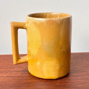 Puede incluir: Una taza de cerámica amarilla con un asa rectangular y un cuerpo cilíndrico. La taza tiene un acabado brillante. La taza está colocada sobre una superficie de madera.