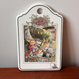 Puede incluir: Placa decorativa Foxwood Tales de cerámica blanca con borde verde. La placa presenta una colorida ilustración de conejos haciendo un picnic. Tiene un agujero en la parte superior para colgar.