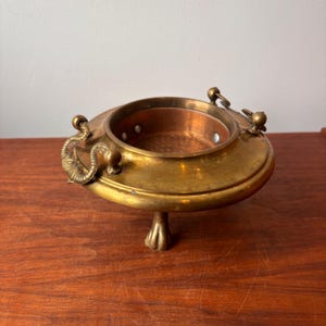 Może przedstawiać: Antyczna mosiężna miska z miedzianym wnętrzem. Miska ma okrągły kształt z ozdobnymi uchwytami i spoczywa na pojedynczej, bogato zdobionej nóżce. Mosiądz ma ciepłą, postarzaną patynę, sugerującą jego wiek. Miska jest prawdopodobnie przeznaczona do celów dekoracyjnych.