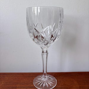 May include: A clear crystal wine glass with a decorative pattern. The glass has a round base, a long stem, and a bowl with a diamond and vertical line design. The glass is designed for serving wine.