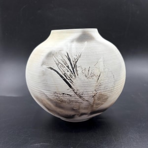 Puede incluir: Un jarrón de cerámica blanca con cuerpo redondo y un diseño abstracto en negro y gris. El jarrón mide aproximadamente 15 cm de alto.