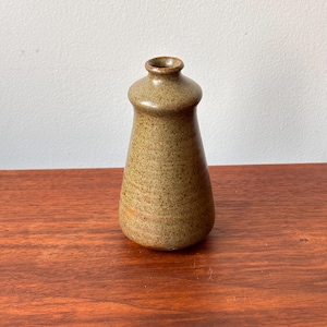 May include: A small, handcrafted ceramic vase with a speckled, earthy brown glaze. The vase has a rounded top and a slightly flared base. The overall shape is reminiscent of a bottle. The vase is displayed on a wooden surface.