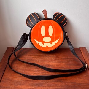 May include: Orange pumpkin-shaped crossbody bag with a smiling face and black Mickey Mouse ears. The bag has a black strap and trim. The pumpkin face is cream-colored. The ears are black with orange stripes. A brown stem is at the top.