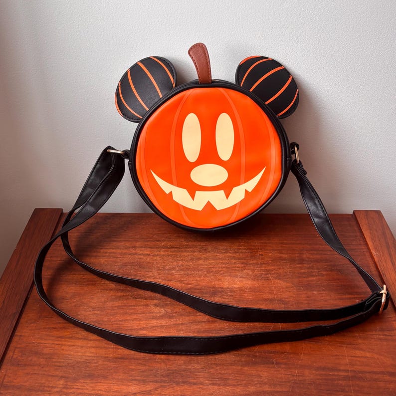 May include: Orange pumpkin-shaped crossbody bag with a smiling face and black Mickey Mouse ears. The bag has a black strap and trim. The pumpkin face is cream-colored. The ears are black with orange stripes. A brown stem is at the top.